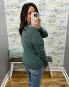 A woman takes a mirror selfie in a fitting room wearing a heathered sage green knit sweater and blue jeans.