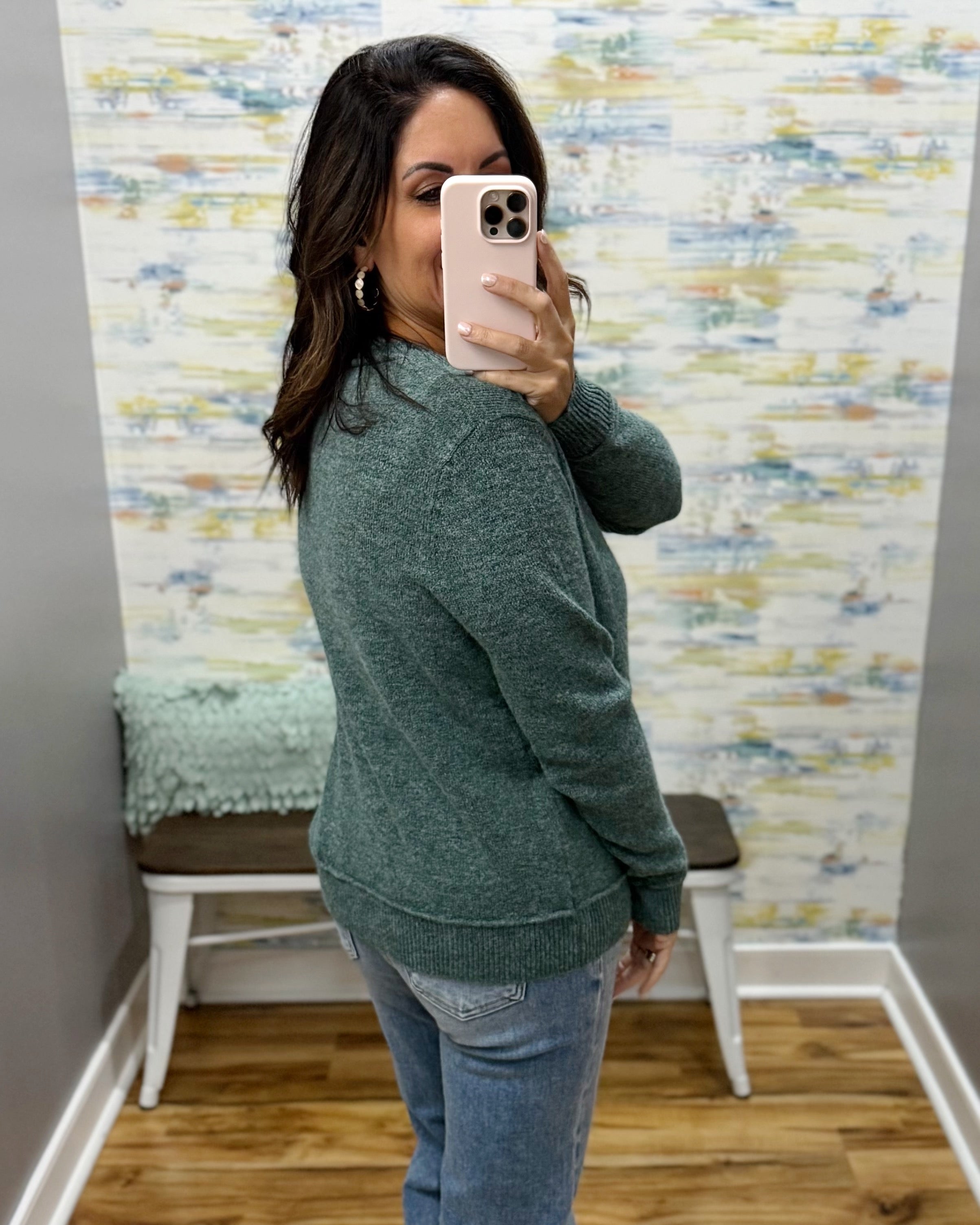 A woman takes a mirror selfie in a fitting room wearing a heathered sage green knit sweater and blue jeans.