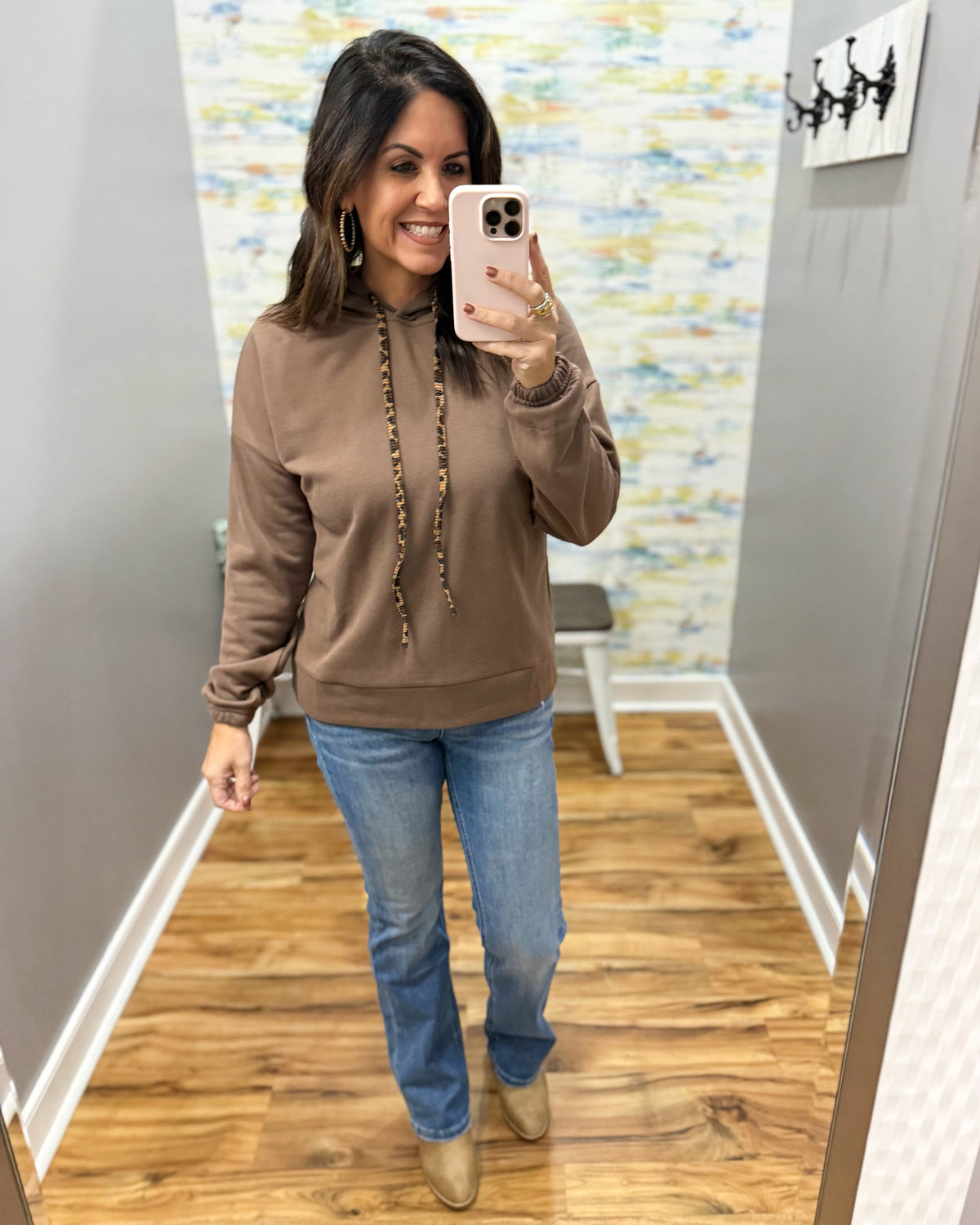 A woman takes a mirror selfie wearing a brown long-sleeve hoodie with a beaded drawstring, mica bootcut jeans, and corkys tan ankle boots in a fitting room with wood floors and gray walls.