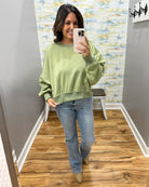 A woman takes a mirror selfie wearing a sage green crewneck sweatshirt and light blue jeans in a fitting room with wood floors and a patterned wall.