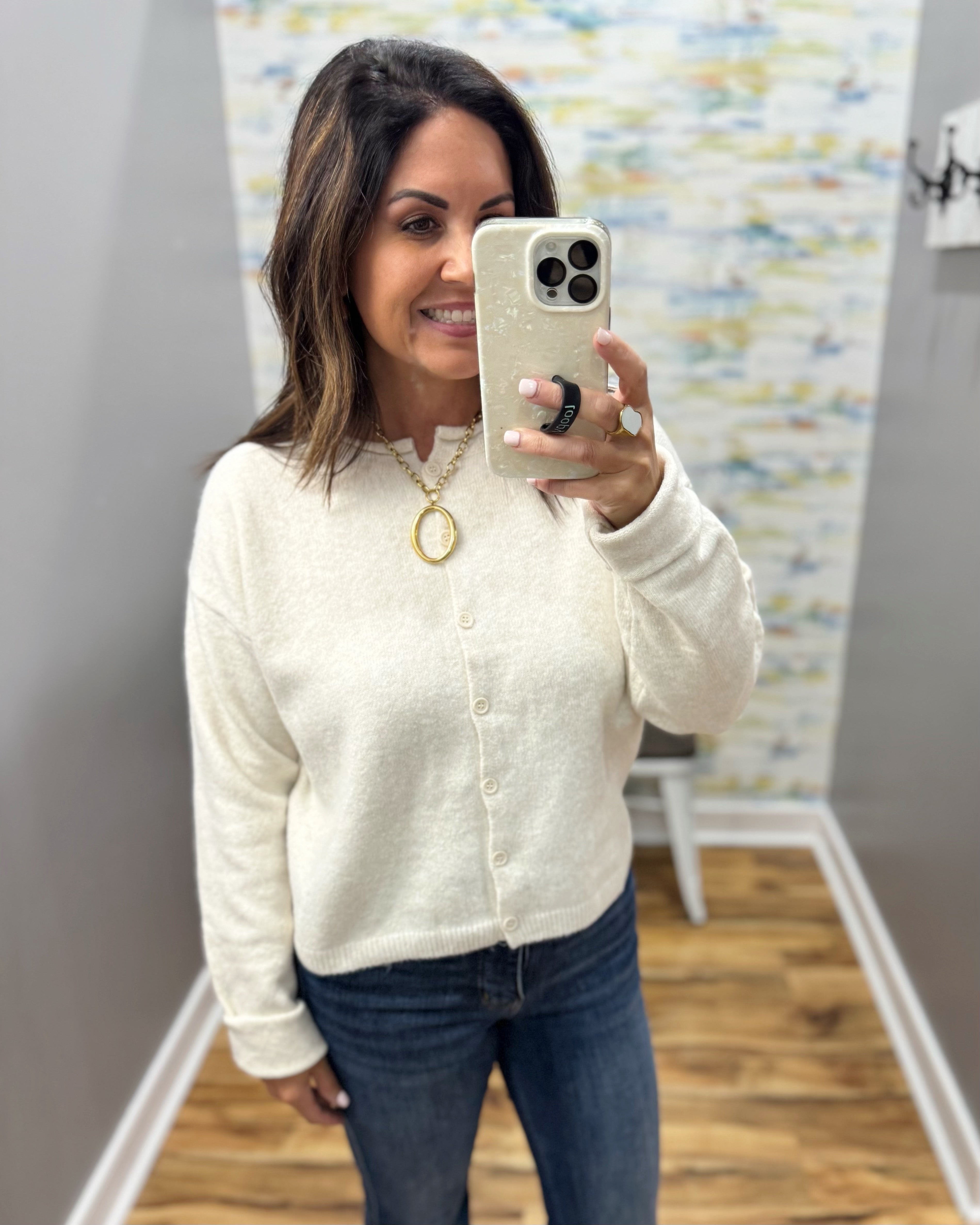 A woman takes a mirror selfie wearing a cream-colored, long-sleeve button-up knit cardigan sweater and dark blue jeans. She is wearing a gold pendant necklace and a black ring on her phone.