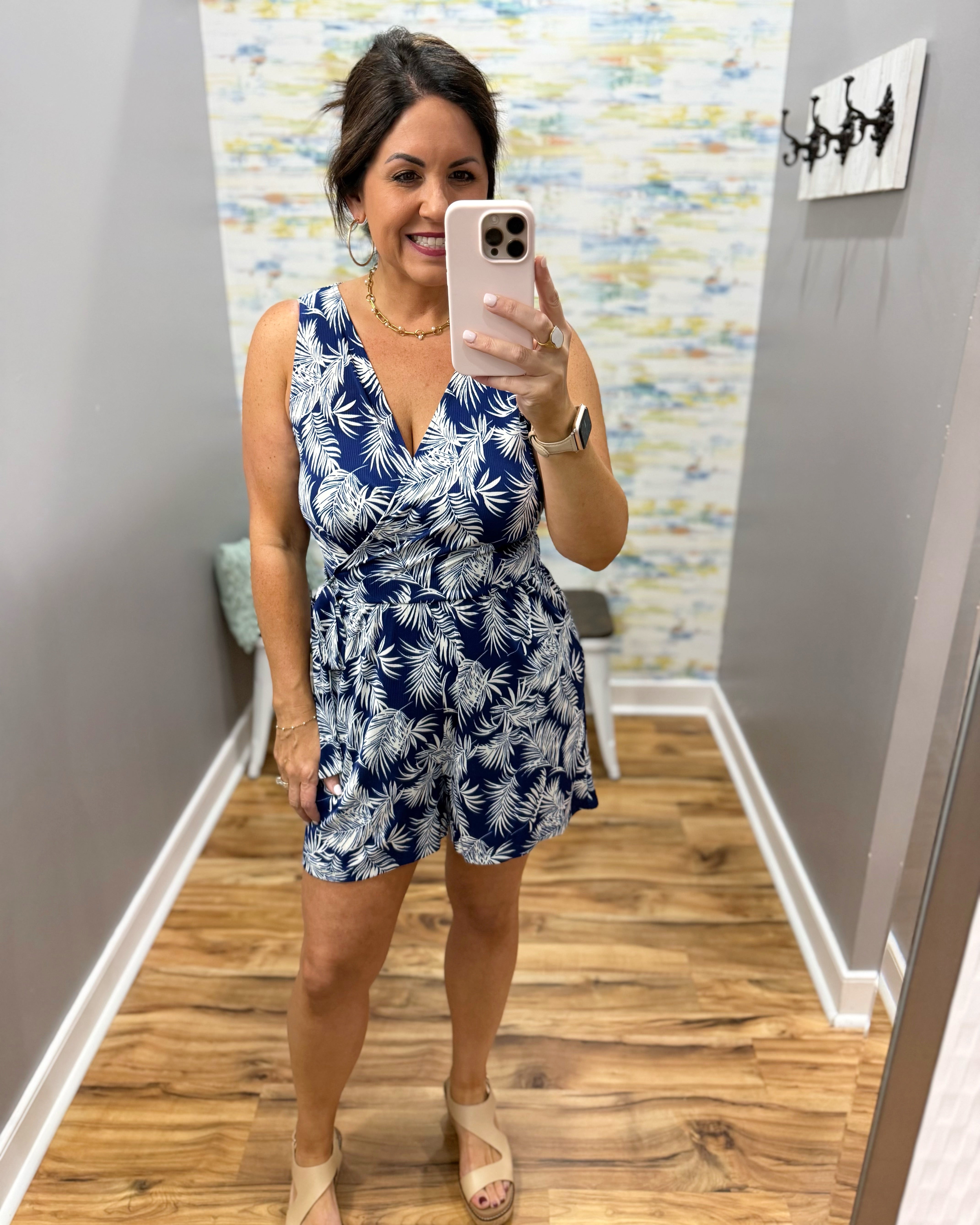 Woman taking a mirror selfie wearing a blue and white patterned romper in a room with wooden flooring and a colorful wall.