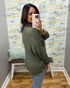 A woman takes a mirror selfie wearing a textured olive green long-sleeve top and light blue jeans in a room with a patterned wallpaper background.