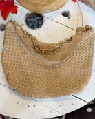A woven tan shoulder bag with a gold chain strap, displayed on a rustic white wooden surface.
