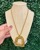 Chansuttpearls gold Taylor necklace with a large abstract square pendant displayed on a beige fabric mannequin bust, with a hand holding the left side of the bust against a green leaf background.