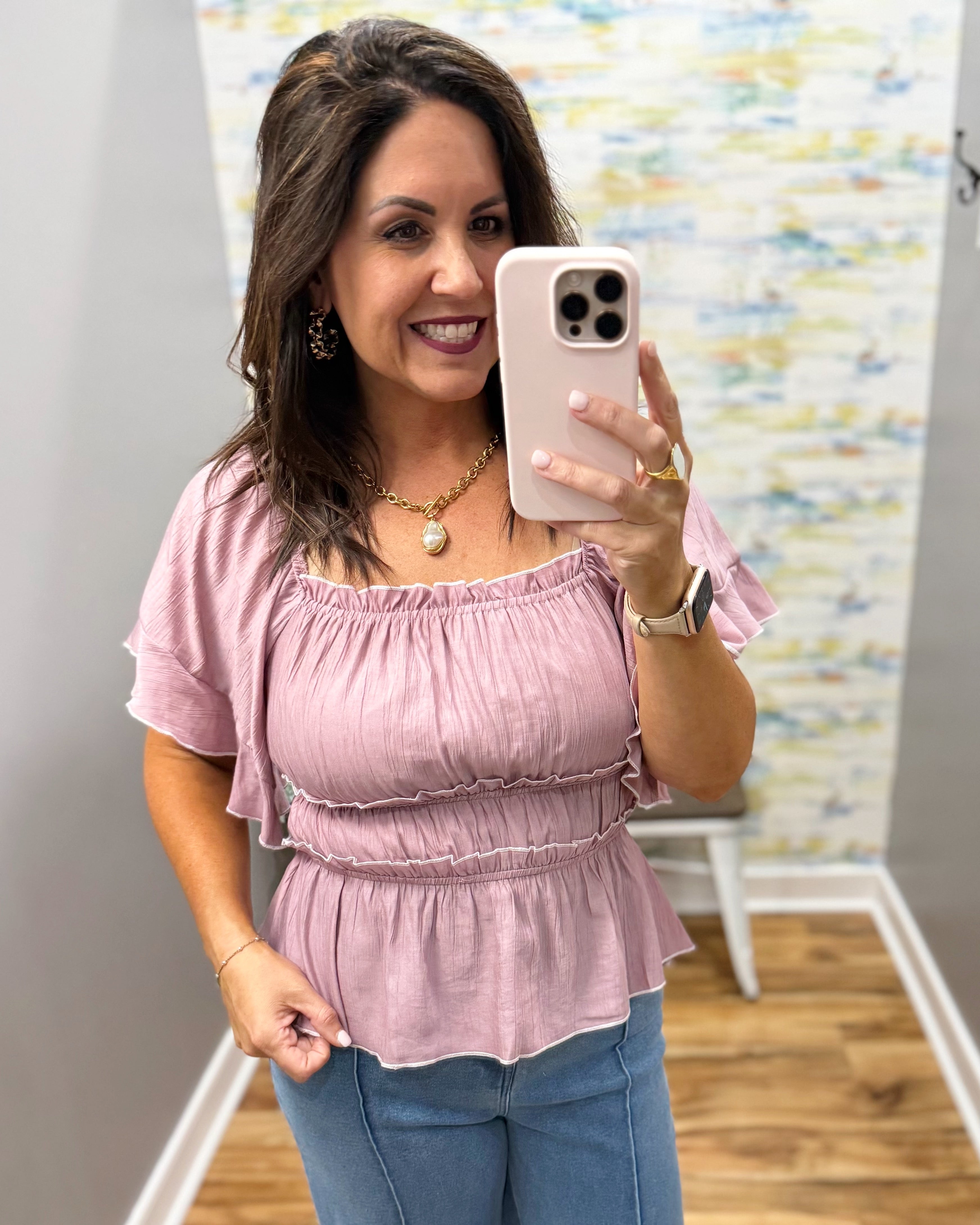 Woman taking a mirror selfie wearing a pink top and blue jeans in a room with colorful wall art.