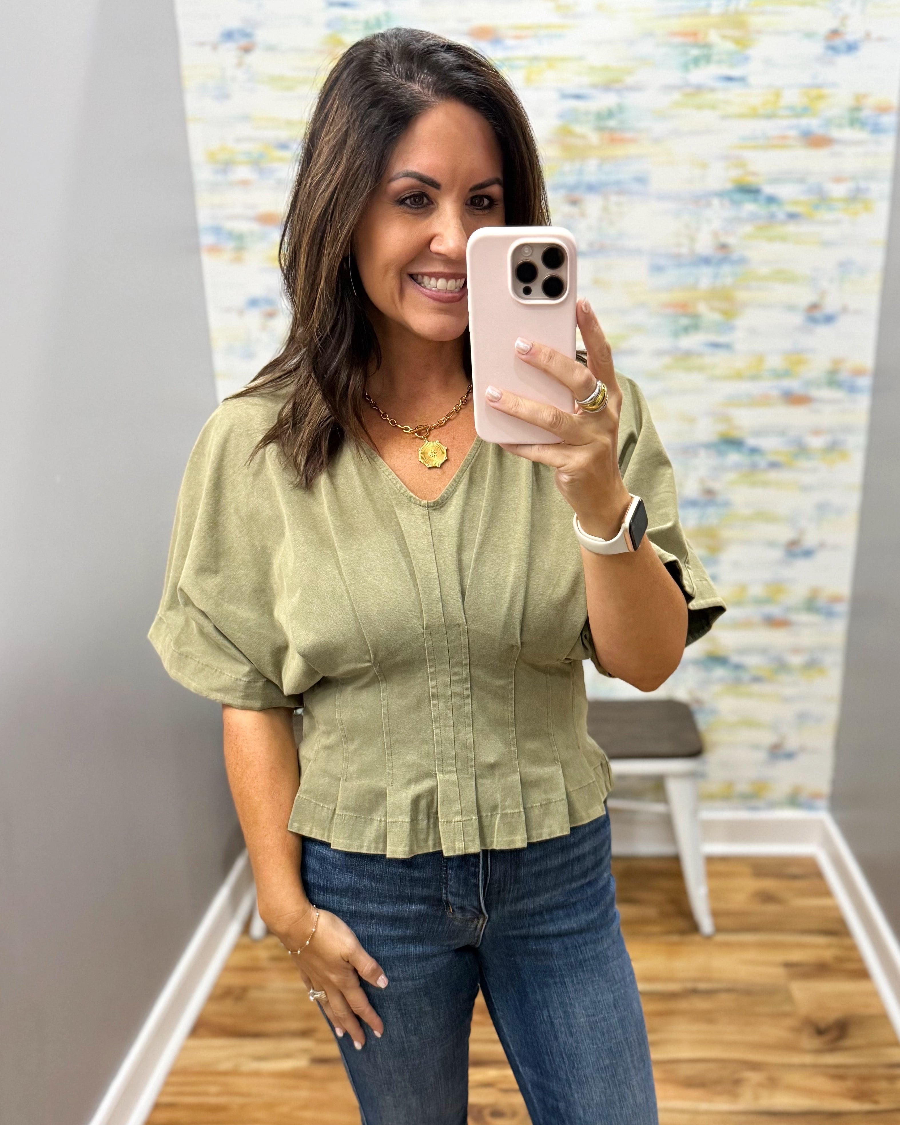 A woman takes a mirror selfie wearing an olive green short-sleeve blouse with puff sleeves and a pleated front, paired with blue jeans.