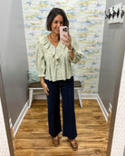 A woman takes a mirror selfie in a fitting room wearing a light sage green ruffled blouse and dark navy wide-leg pants.