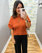 A woman takes a mirror selfie wearing a short-sleeve knit top in burnt orange with black wide-leg pants. She is wearing gold jewelry, including a gold pendant necklace, hoop earrings, and rings, and has a white smartwatch on her wrist.