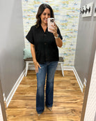 A woman takes a mirror selfie wearing a black short-sleeve button-front top with front pockets and blue flare jeans, standing in a room with wood floors and a patterned wall.