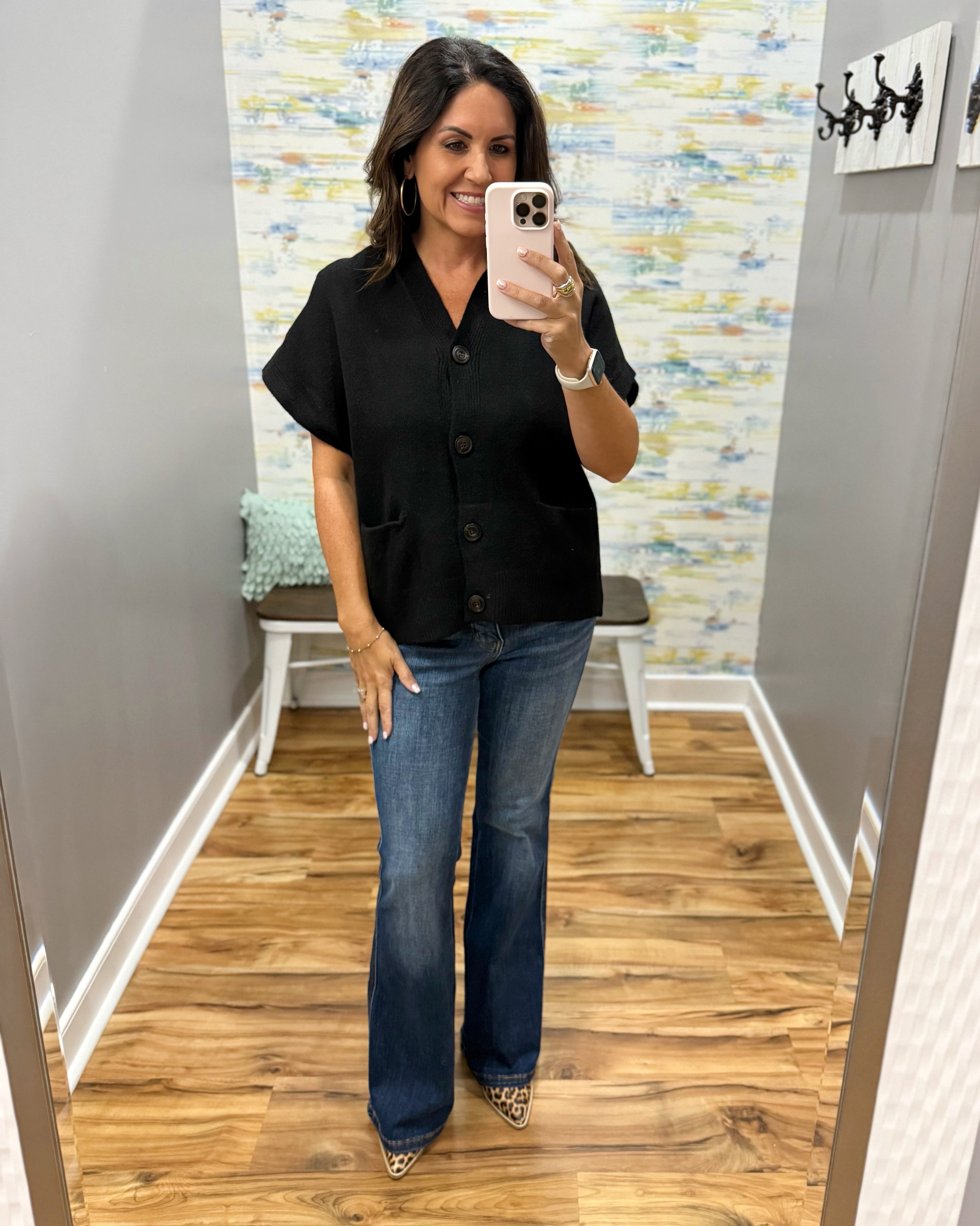 A woman takes a mirror selfie wearing a black short-sleeve button-front top with front pockets and blue flare jeans, standing in a room with wood floors and a patterned wall.