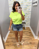 Woman taking a mirror selfie wearing a bright green t-shirt and denim shorts in a room with a patterned wall.