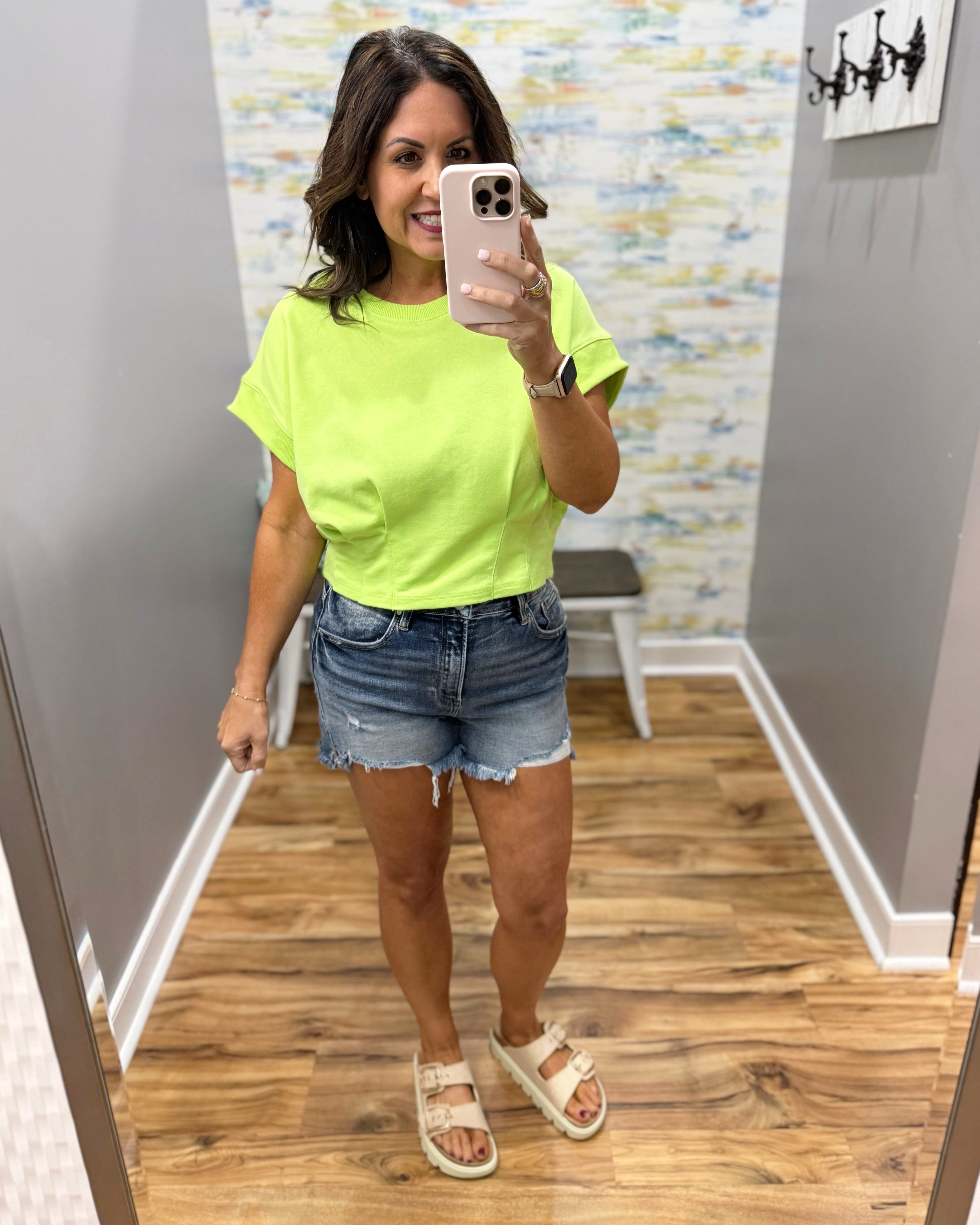 Woman taking a mirror selfie wearing a bright green t-shirt and denim shorts in a room with a patterned wall.