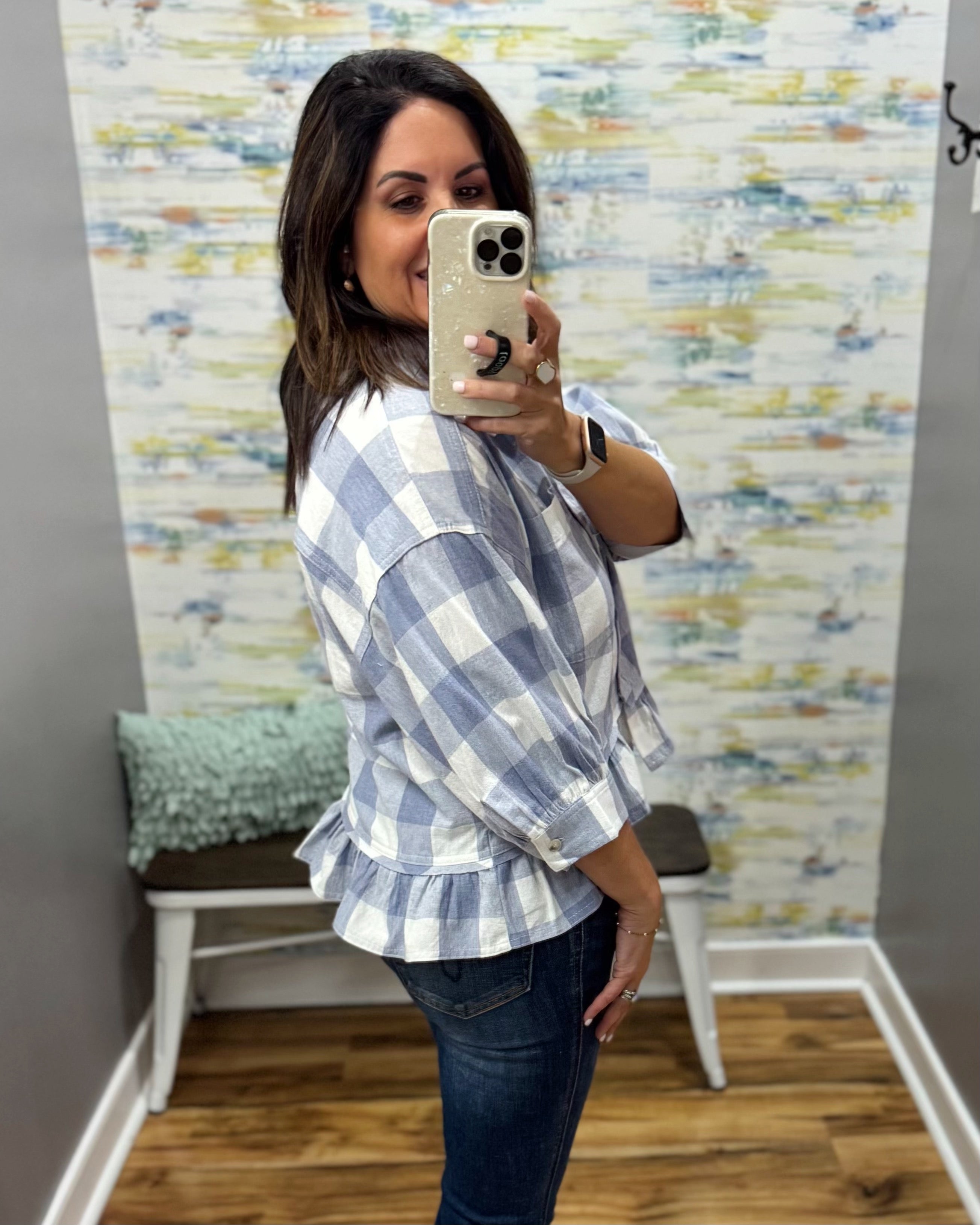 A woman takes a mirror selfie wearing a light blue and white gingham ruffled blouse with dark blue jeans in a fitting room with patterned wallpaper.
