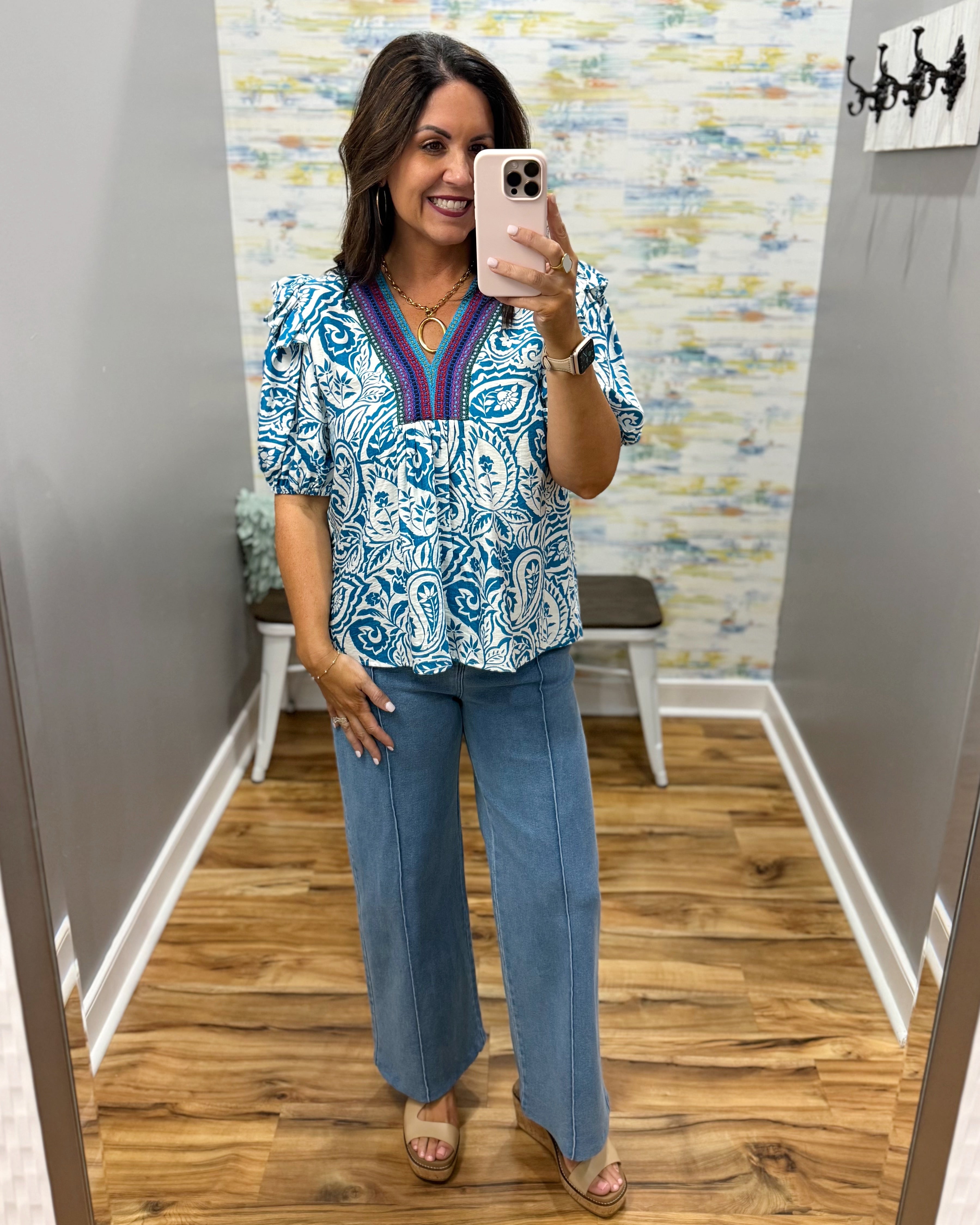 Woman taking a mirror selfie wearing a patterned top and blue jeans in a room with a colorful wall.