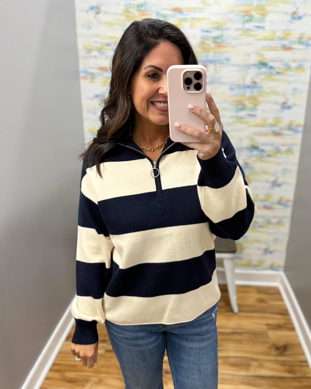 A woman taking a mirror selfie in a boutique, wearing a navy blue and cream horizontal striped quarter-zip pullover sweater with blue jeans.