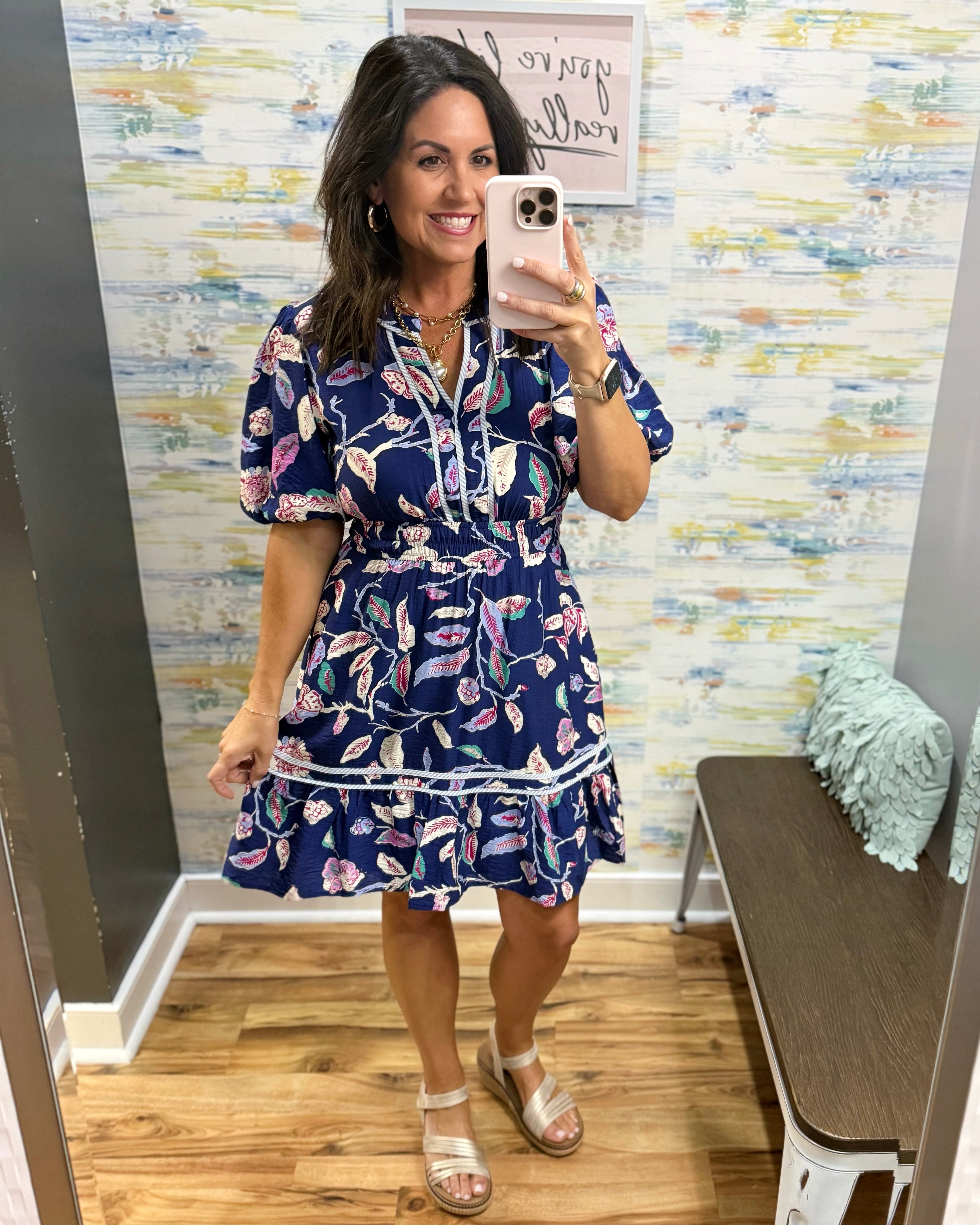 Woman taking a mirror selfie wearing a blue floral dress in a room with colorful wall art.