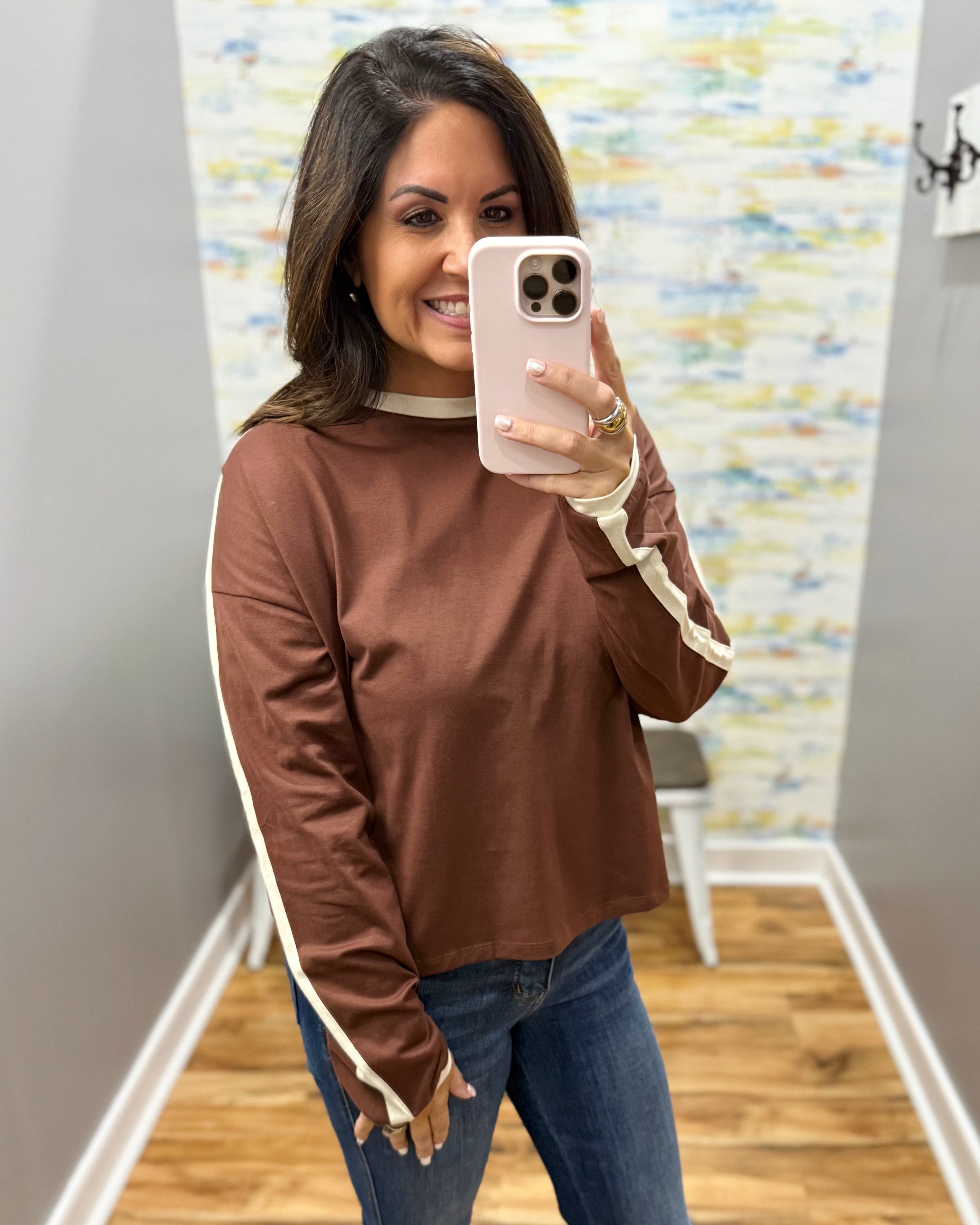 A woman taking a mirror selfie in a room wearing a brown long-sleeve top with beige stripes down the sleeves and a beige crew neck trim, paired with blue jeans.