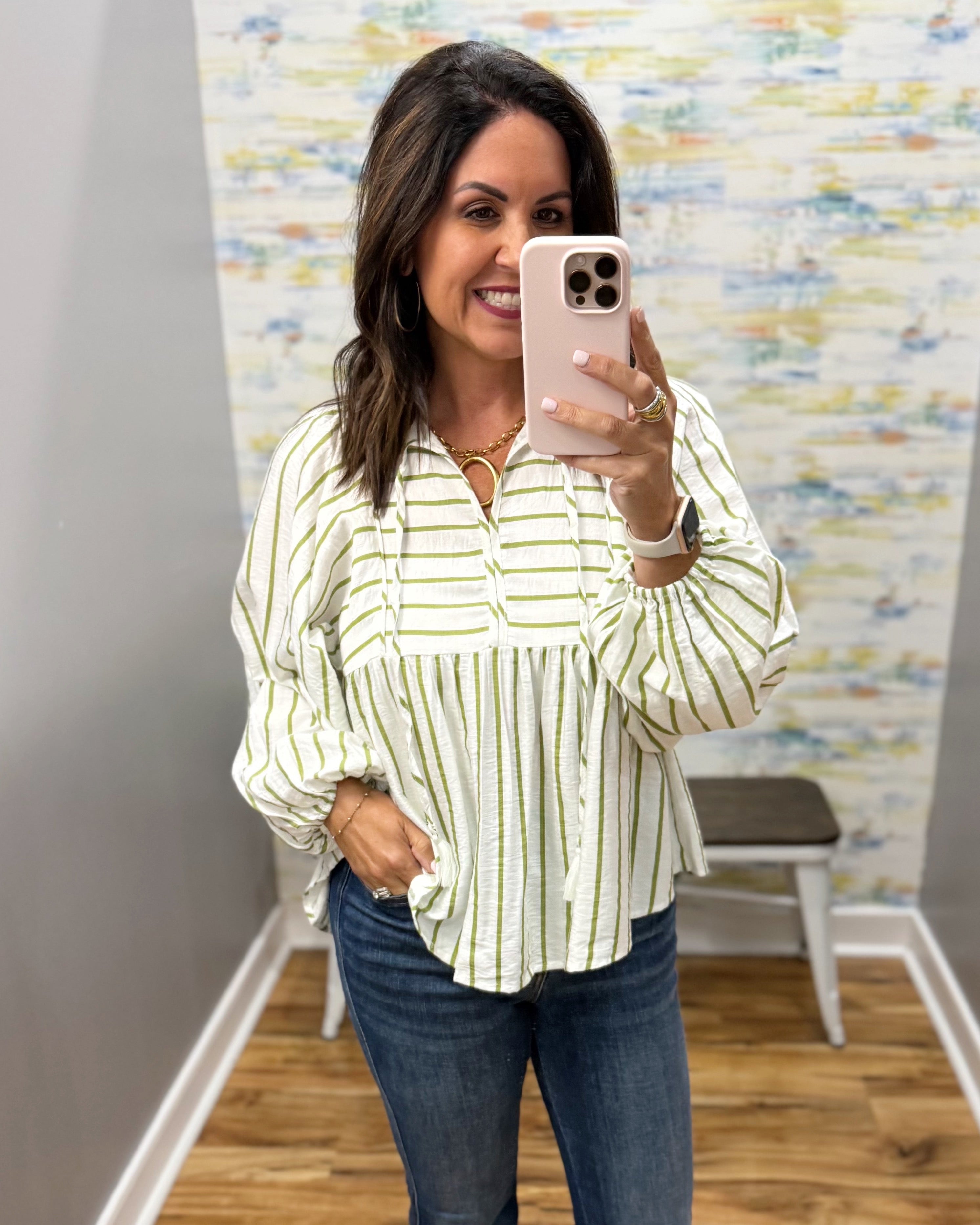 A woman takes a mirror selfie wearing a cream and green vertically striped blouse with blue jeans, standing in front of a patterned wall.