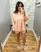 A woman takes a mirror selfie in a fitting room wearing a peach and white striped long-sleeve t-shirt and matching shorts, paired with beige slide sandals.