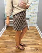 A person wearing a beige knit sweater and a brown and beige argyle skirt, paired with Corkys Pity Party in Cognac wedge sandals, standing in a room with wood floors and a mirror selfie.