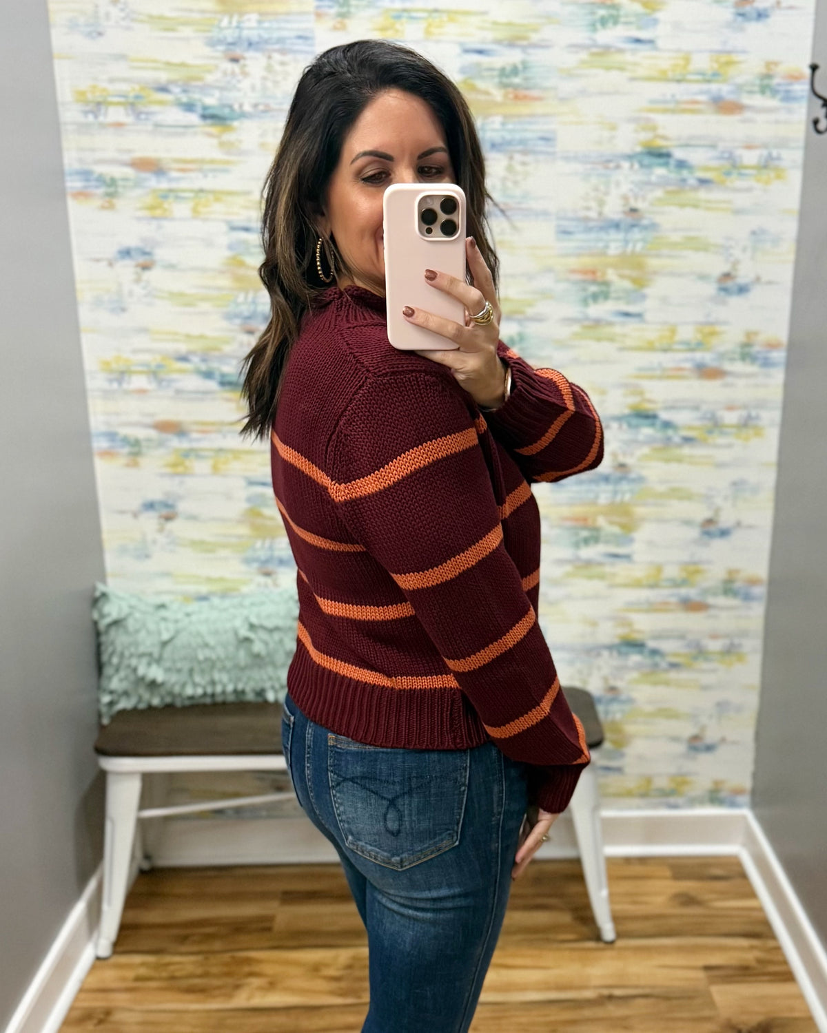 A woman takes a mirror selfie wearing a burgundy knit sweater with orange horizontal stripes and blue jeans, standing in front of a patterned wallpaper background.