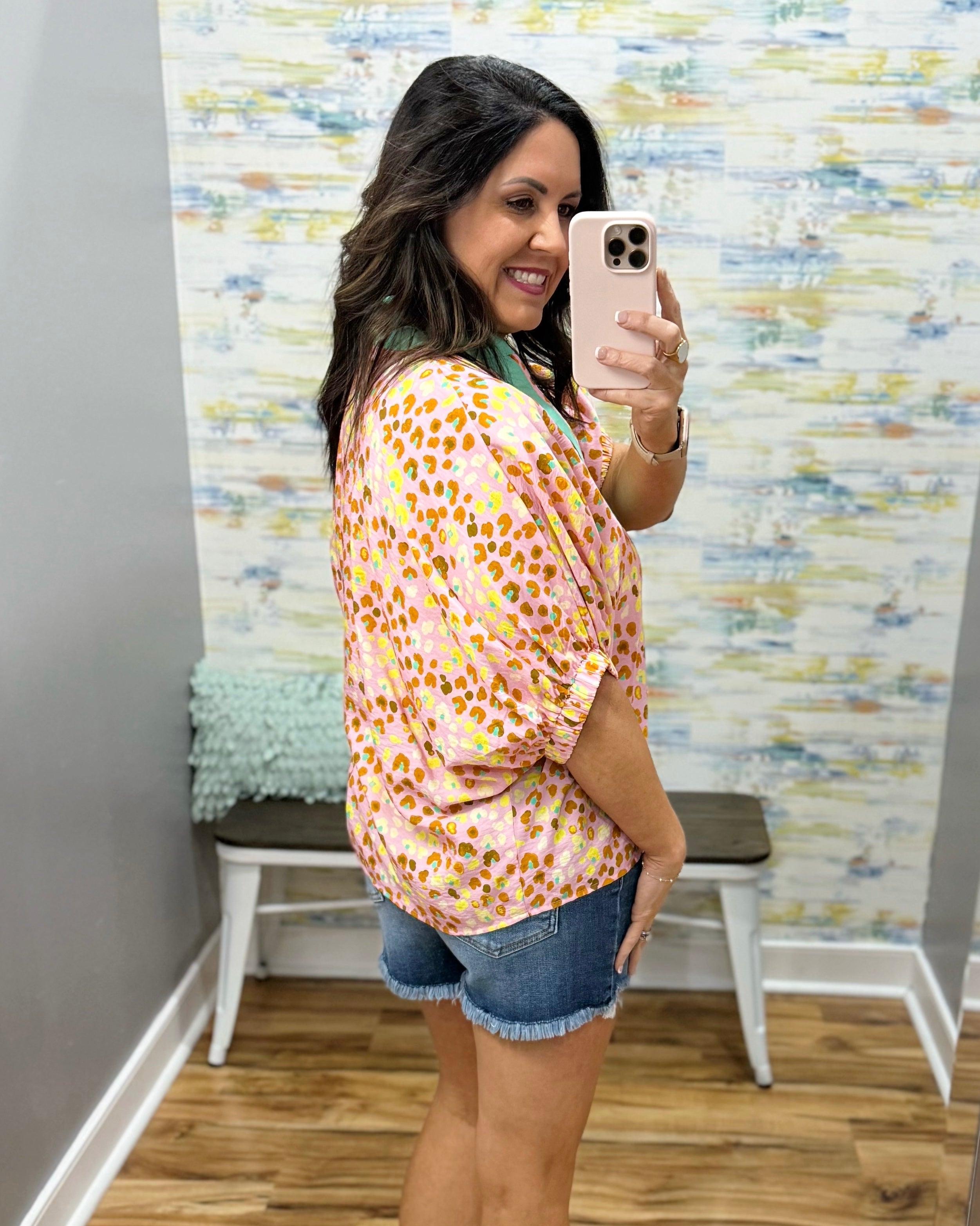 Woman taking a mirror selfie wearing a floral blouse and denim shorts in a room with a textured wall.