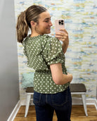 A woman takes a mirror selfie in a fitting room wearing an olive green short-sleeve blouse with white polka dots and dark blue jeans.