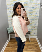 A woman takes a mirror selfie wearing a knit sweater vest with a cream and light pink color-blocked design, paired with dark blue jeans.