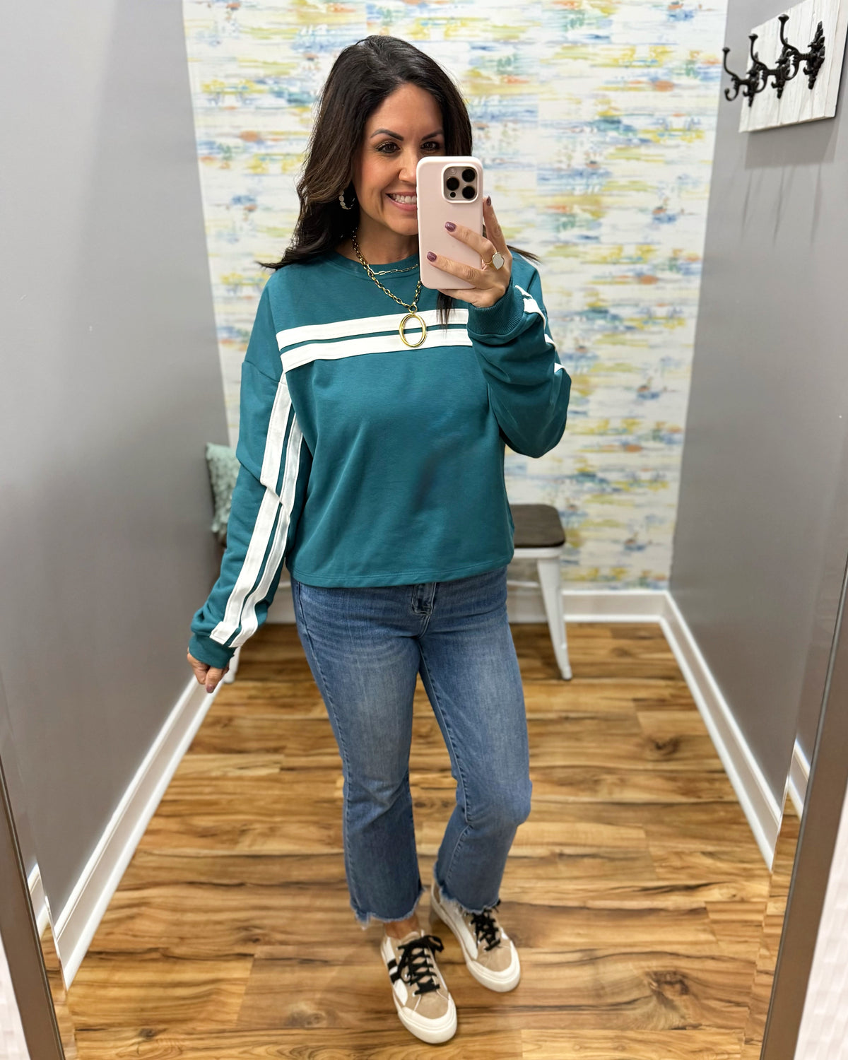A woman takes a mirror selfie wearing a teal long-sleeve sweatshirt with white stripes down the sleeves, paired with blue jeans and beige sneakers, in a fitting room.