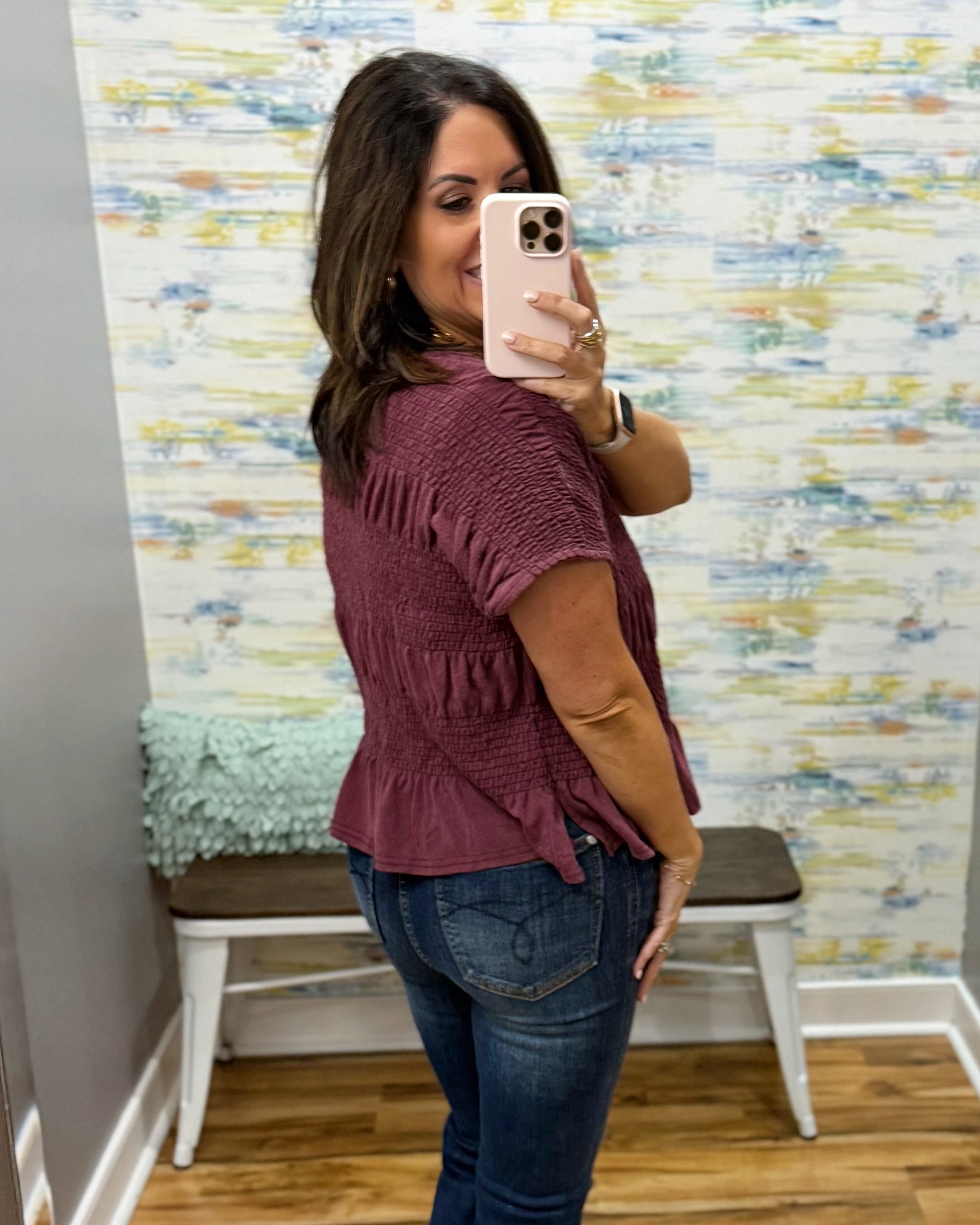 A woman takes a mirror selfie in a fitting room wearing a short-sleeve smocked blouse in a dusty rose color with dark wash jeans.
