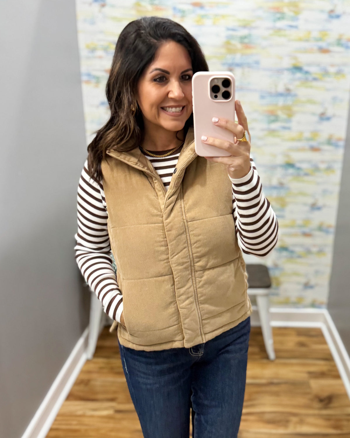 A woman takes a mirror selfie wearing a tan quilted puffer vest over a black and white striped long-sleeve shirt, dark blue jeans, and tan boots in a home fitting room.