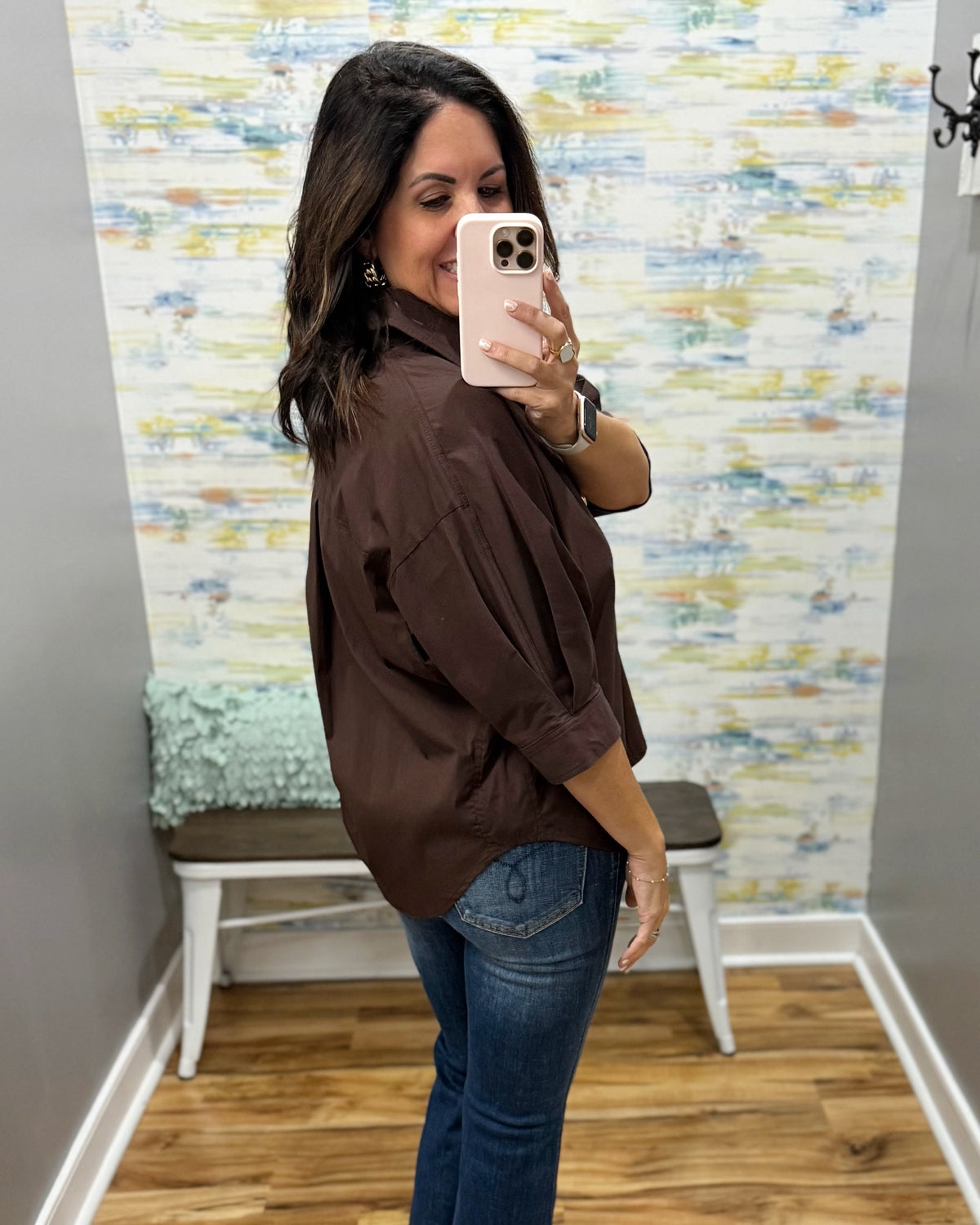 A woman takes a mirror selfie in a fitting room wearing an oversized dark brown button-up shirt and blue jeans.