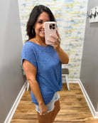 Woman taking a mirror selfie wearing a blue top and white shorts in a room with patterned wallpaper.