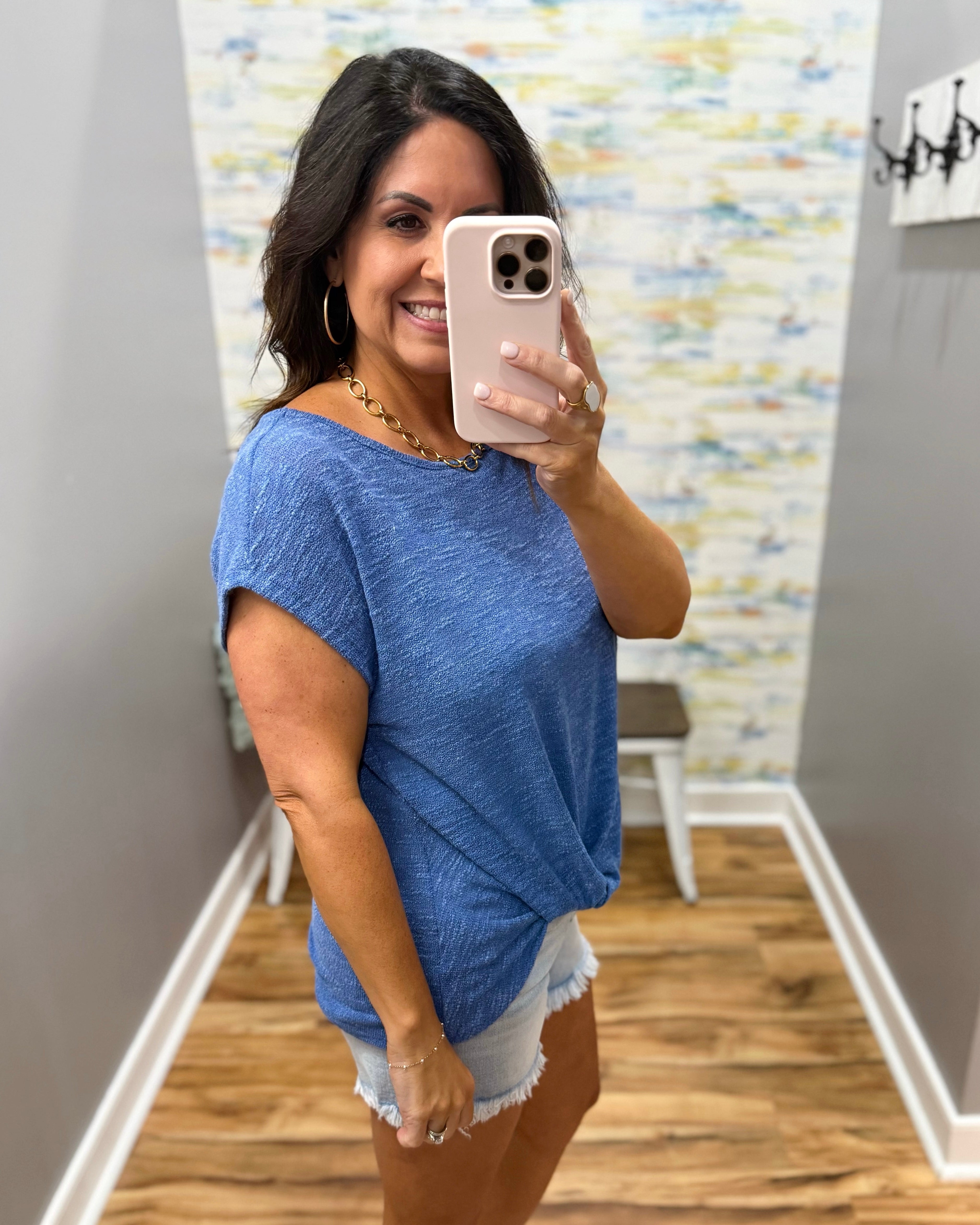 Woman taking a mirror selfie wearing a blue top and white shorts in a room with patterned wallpaper.