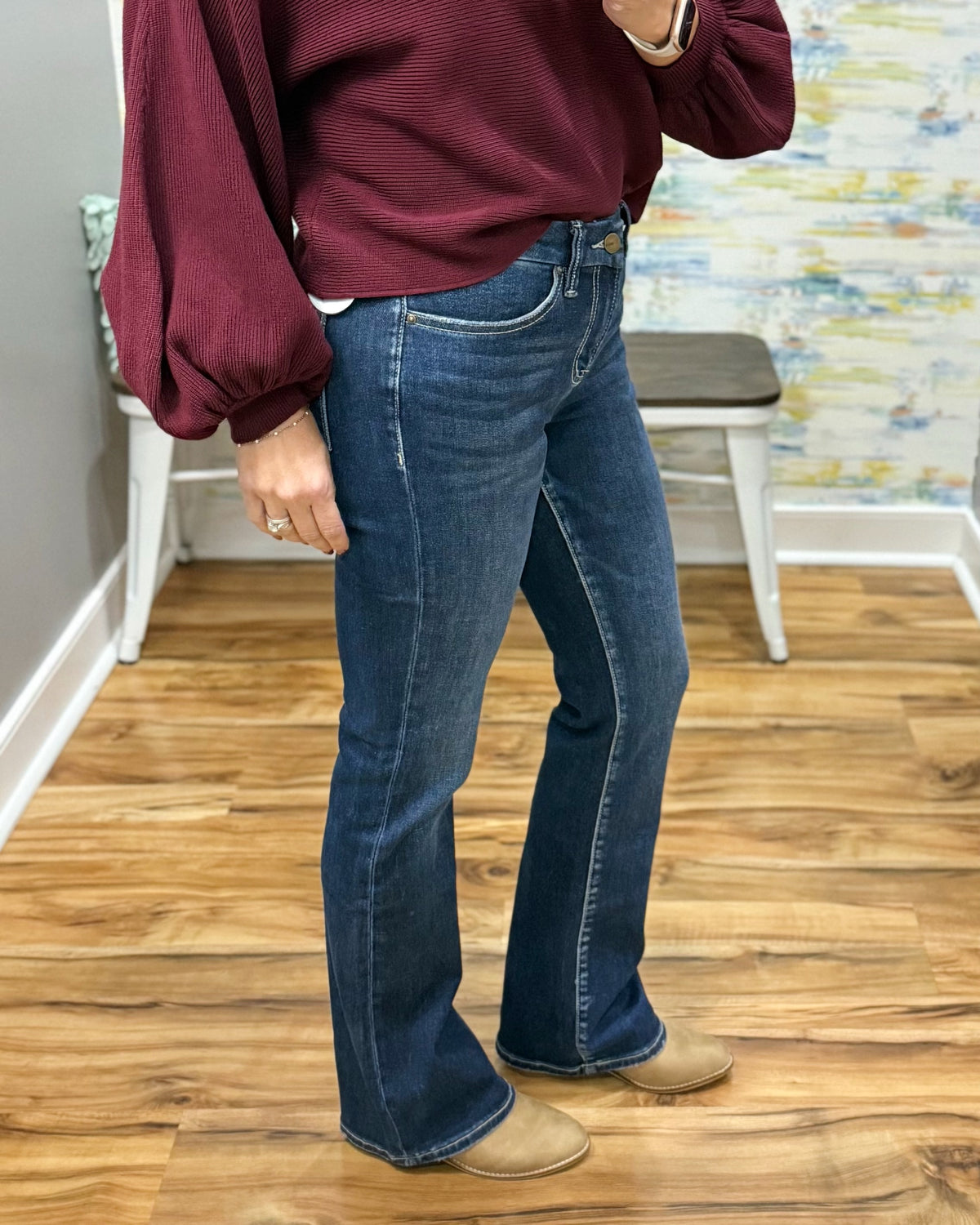 A person wearing mica dark blue flare jeans, a burgundy long-sleeve top, and tan slip-on shoes, standing on a wood floor in an indoor room with a chair and textured wall in the background.