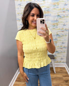 A woman takes a mirror selfie wearing a light yellow short-sleeve smocked peplum top and blue jeans in an indoor setting.