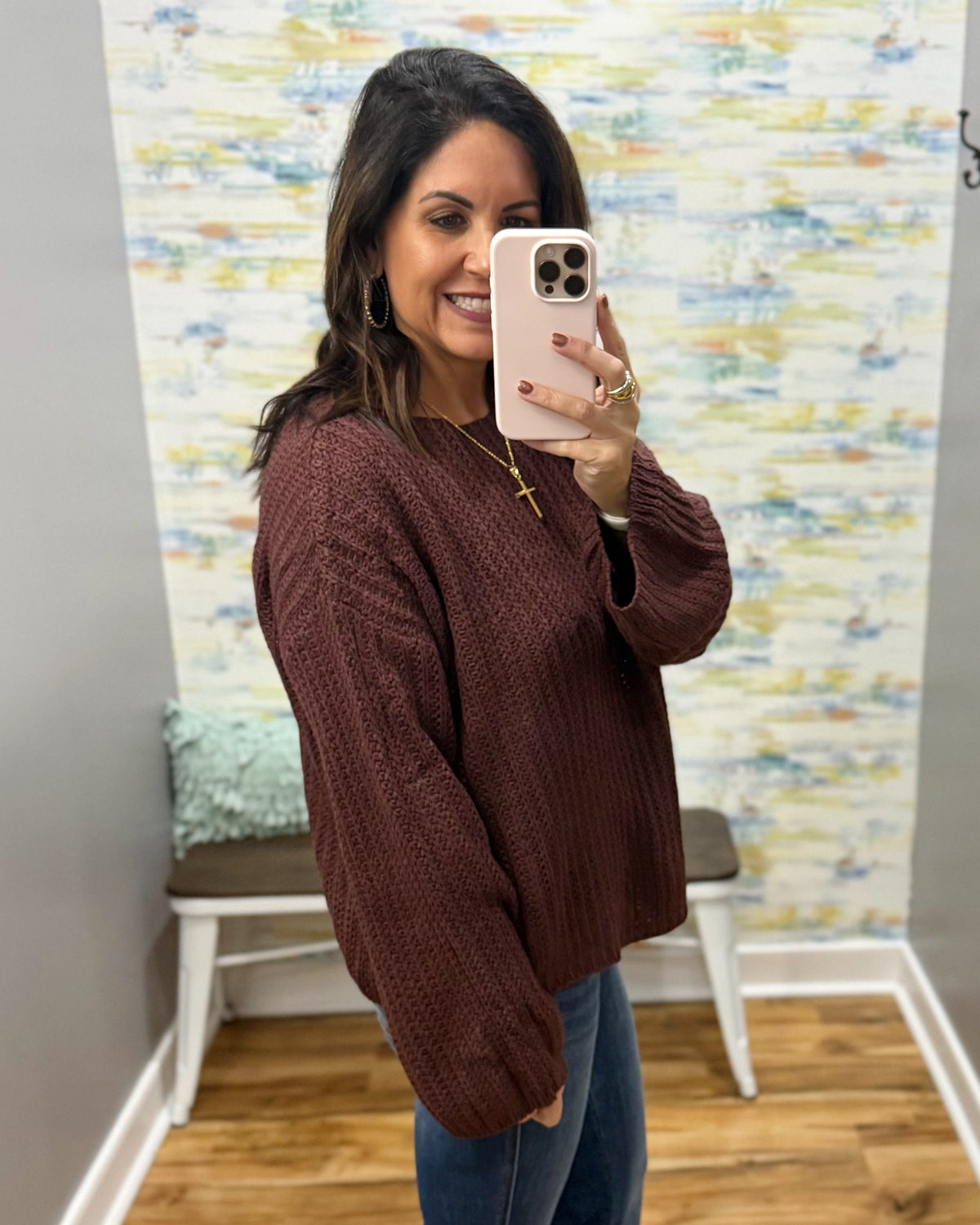 A woman takes a mirror selfie wearing a ribbed knit long-sleeve sweater in a deep burgundy brown color, paired with blue jeans. The setting is a fitting room with a patterned wallpaper background and a wooden floor.