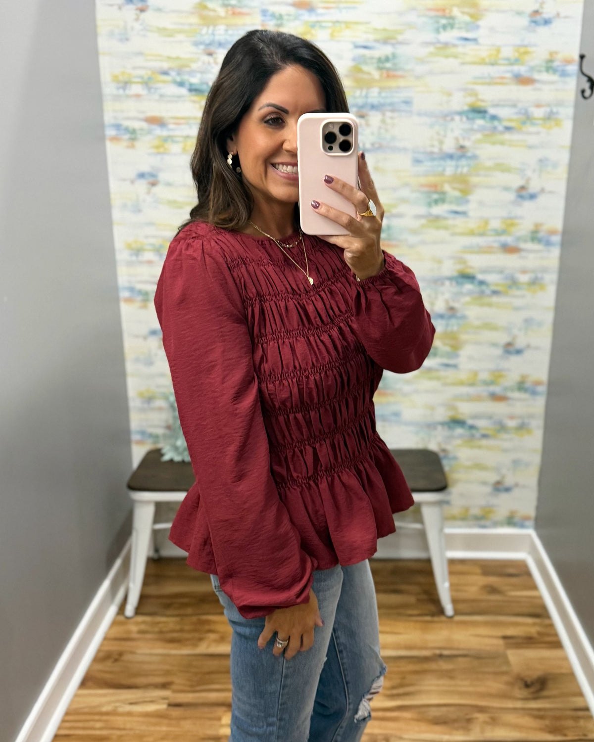 A woman taking a mirror selfie wearing a long-sleeve burgundy smocked blouse and blue jeans in a fitting room or boutique.