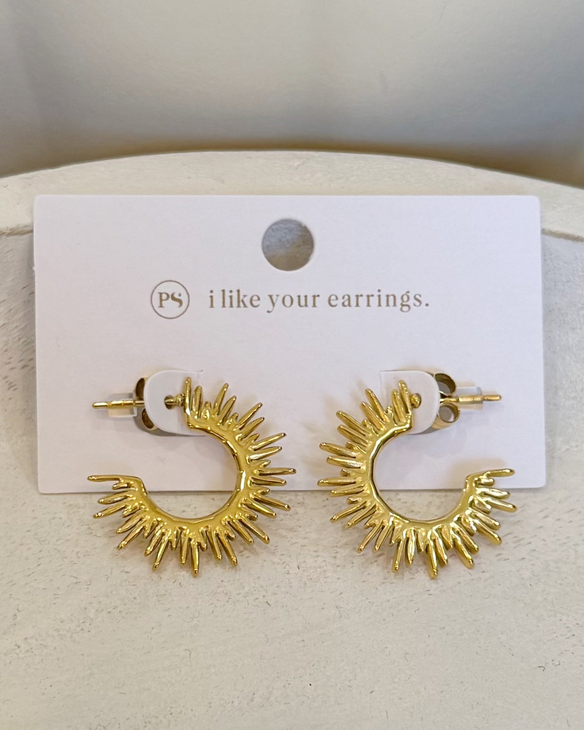 A pair of gold crescent-shaped hoop earrings with radiating sunburst spikes, displayed on a white card that reads 'i like your earrings.' with a small 'ps' logo.