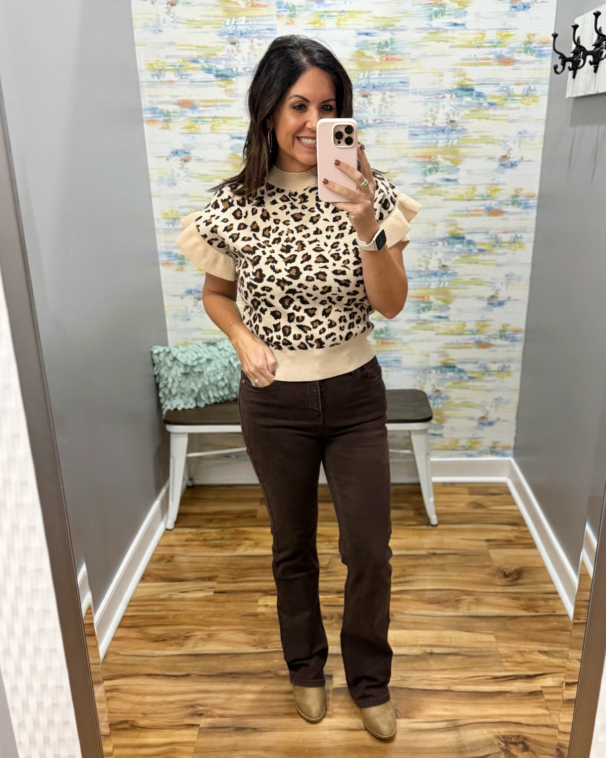 A woman takes a mirror selfie in a fitting room wearing a beige and black leopard print short-sleeve top with ruffle details, paired with dark brown pants and tan shoes.