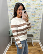 A woman takes a mirror selfie wearing a cream and brown horizontally striped knit sweater with blue jeans, holding a light pink smartphone.