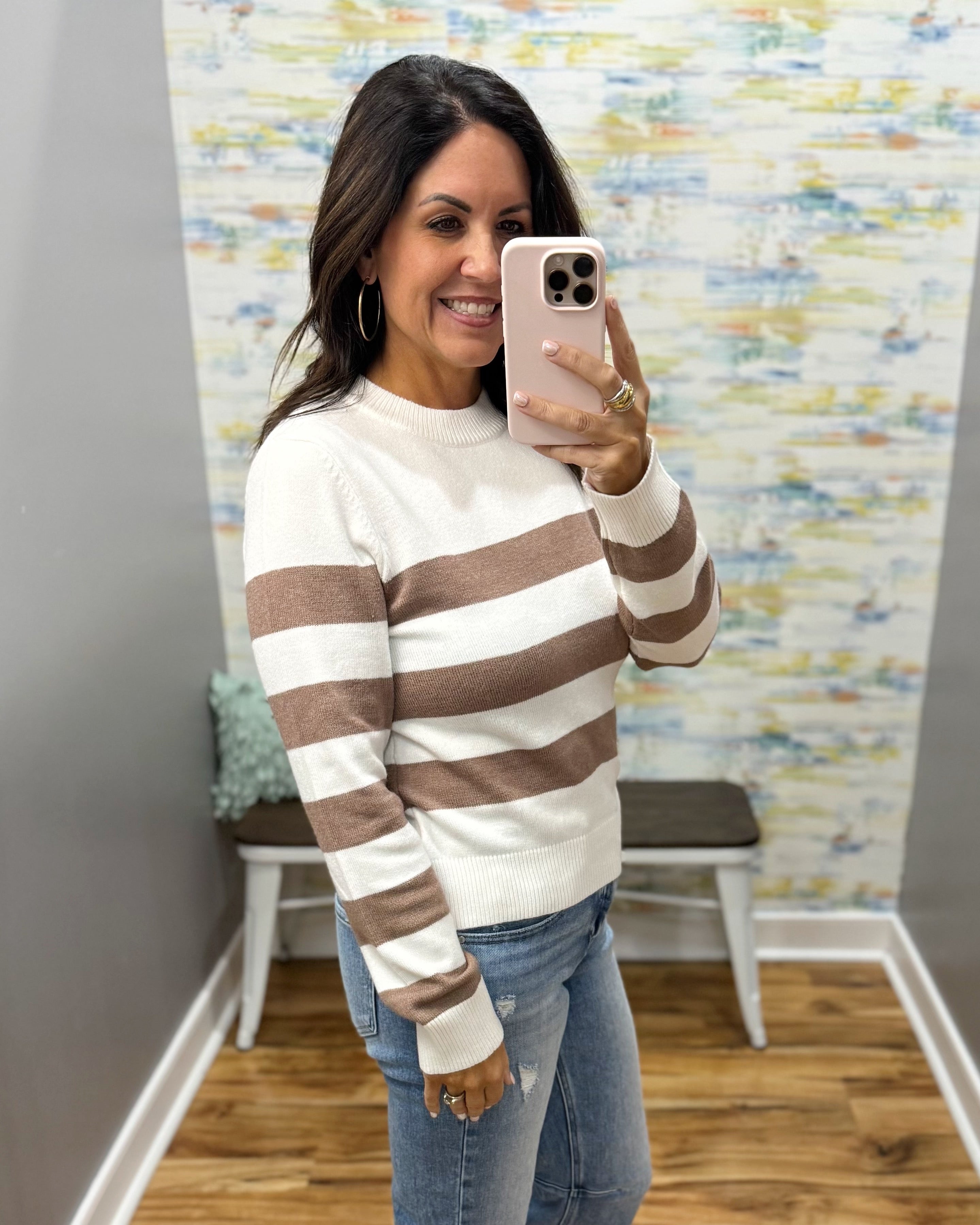 A woman takes a mirror selfie wearing a cream and brown horizontally striped knit sweater with blue jeans, holding a light pink smartphone.