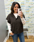 A woman takes a mirror selfie wearing a dark brown knit button-front vest over a pink and white striped long-sleeve shirt, paired with blue jeans. The background shows a gray wall, a wooden floor, and a colorful abstract-patterned wallpaper.