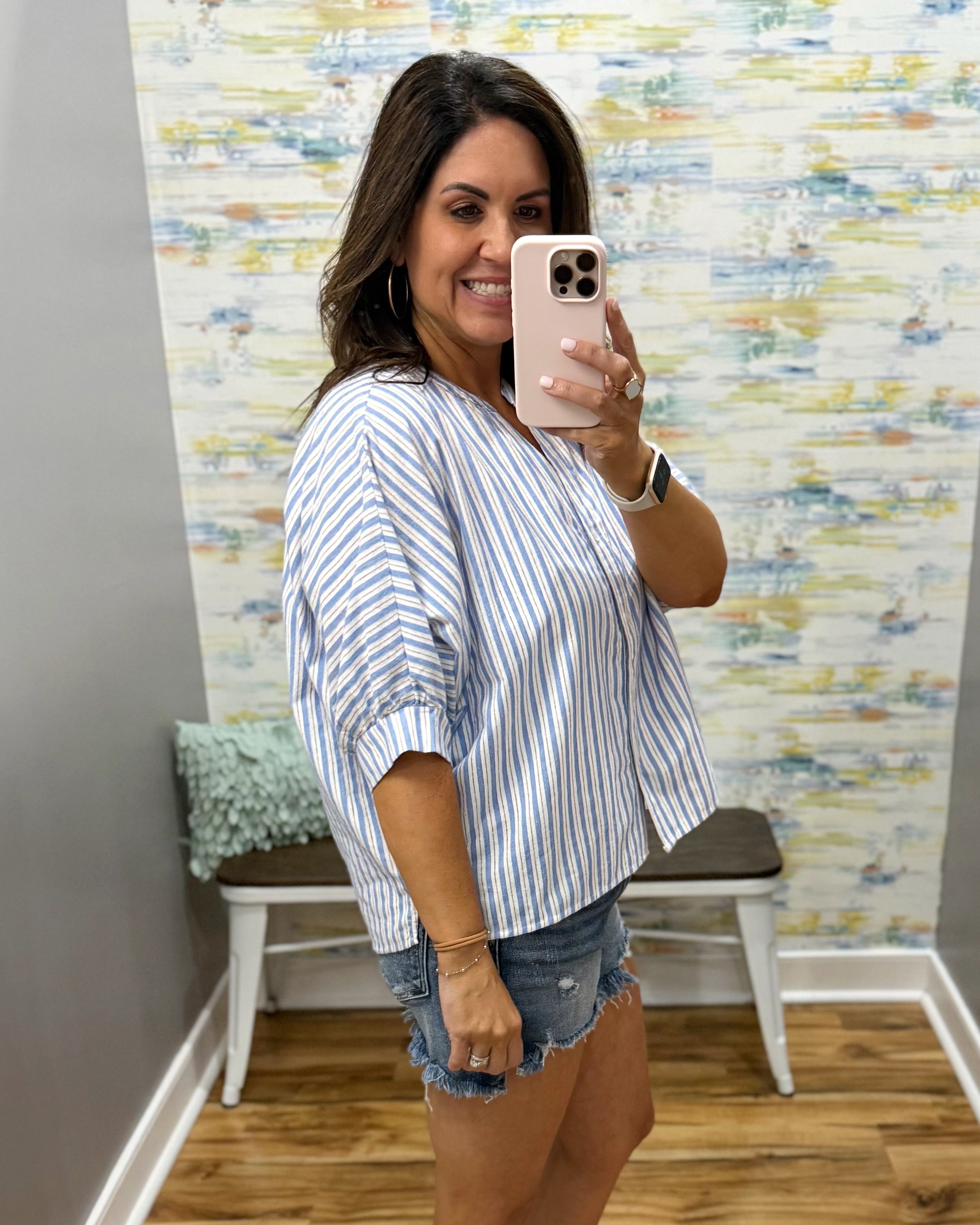 Woman taking a mirror selfie wearing a striped shirt and denim shorts in a room with a textured wall.