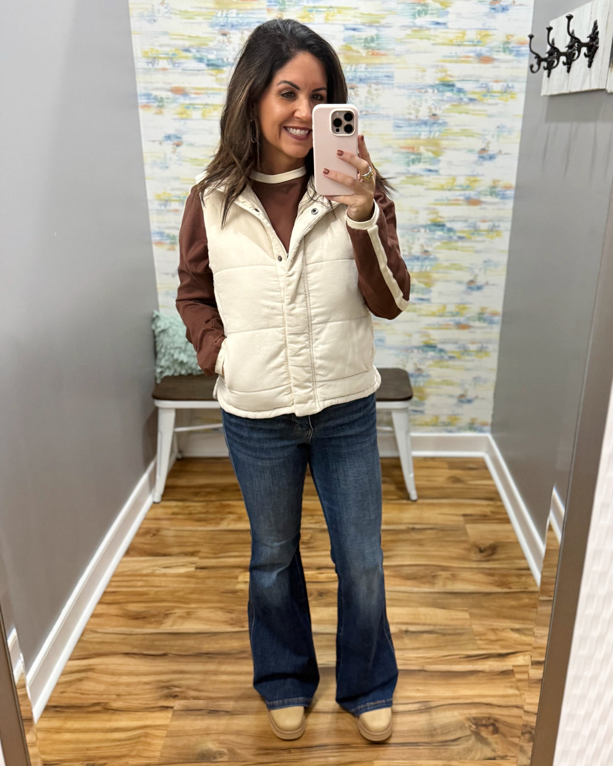 A woman takes a mirror selfie in a fitting room wearing a cream puffer vest over a brown long-sleeve top, blue flare jeans, and tan boots.
