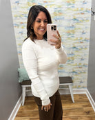 A woman taking a mirror selfie in a fitting room wearing a white ribbed long-sleeve knit sweater and brown pants.