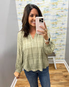 A woman takes a mirror selfie wearing a sage green and beige plaid long-sleeve blouse with blue jeans in an indoor setting with a patterned wallpaper background.