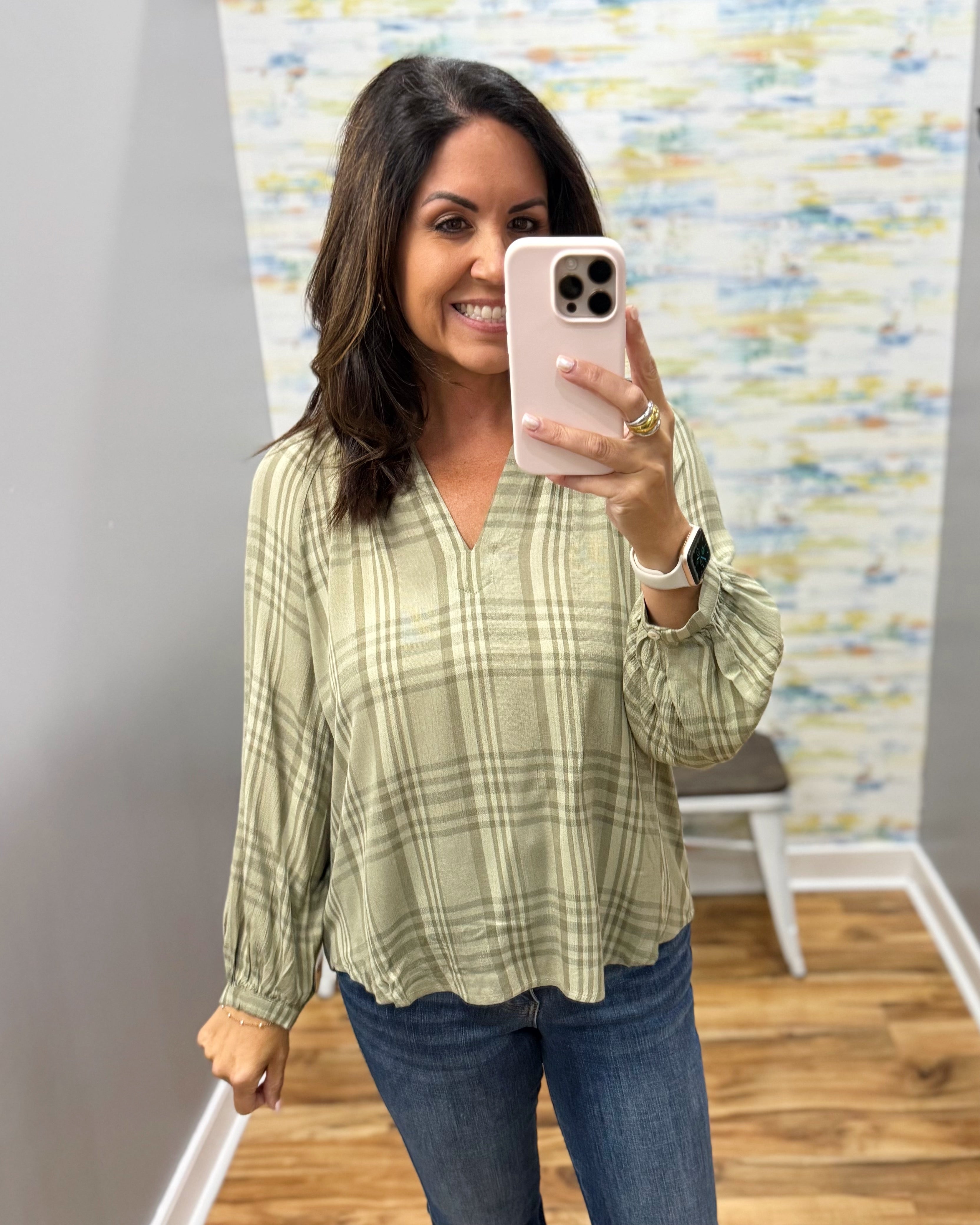 A woman takes a mirror selfie wearing a sage green and beige plaid long-sleeve blouse with blue jeans in an indoor setting with a patterned wallpaper background.