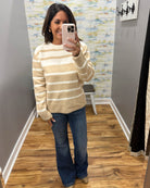A woman takes a mirror selfie in a fitting room wearing a beige and white striped knit sweater, blue jeans, and tan boots.